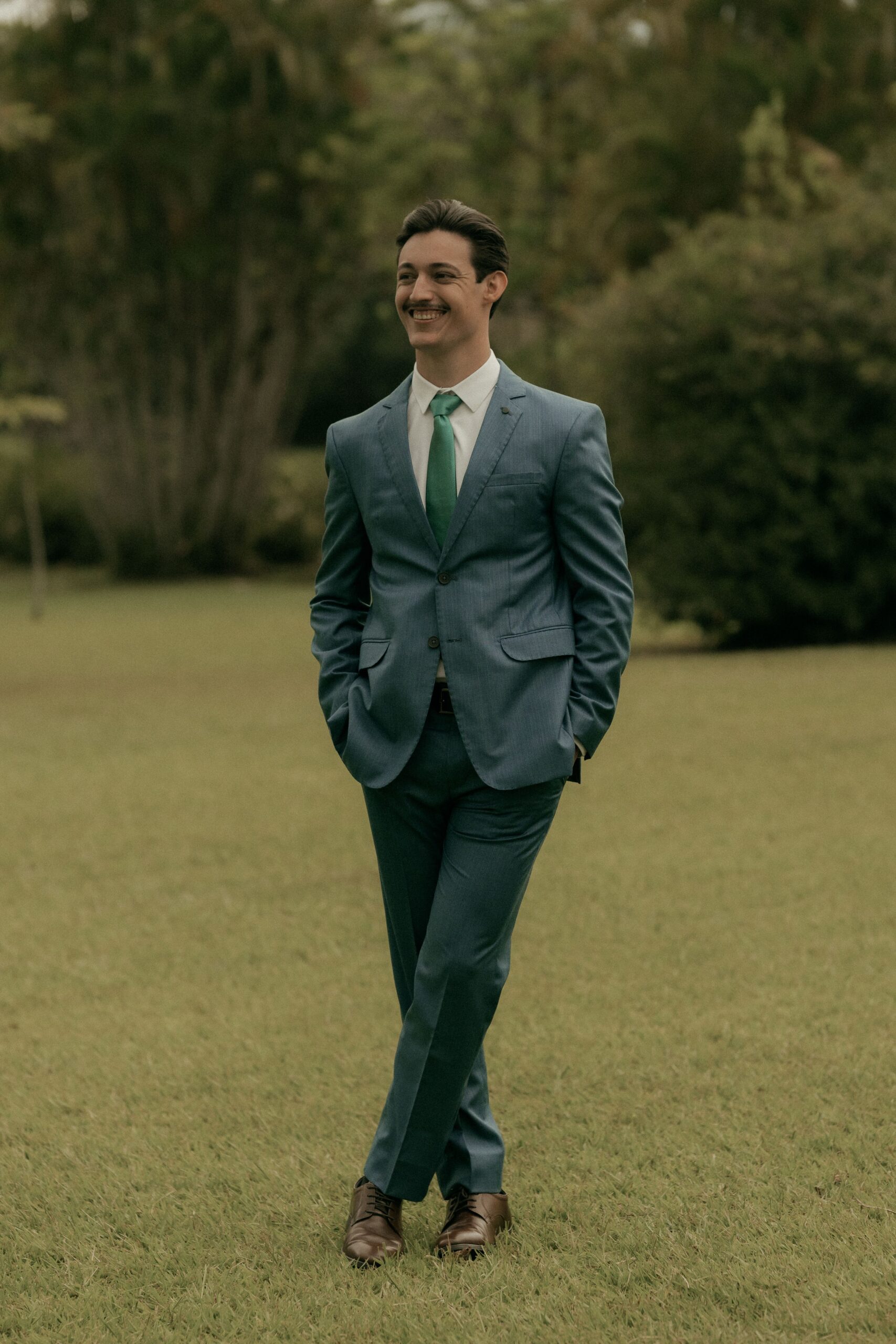 Smiling man in a blue suit with a green tie standing on grass in a park setting.